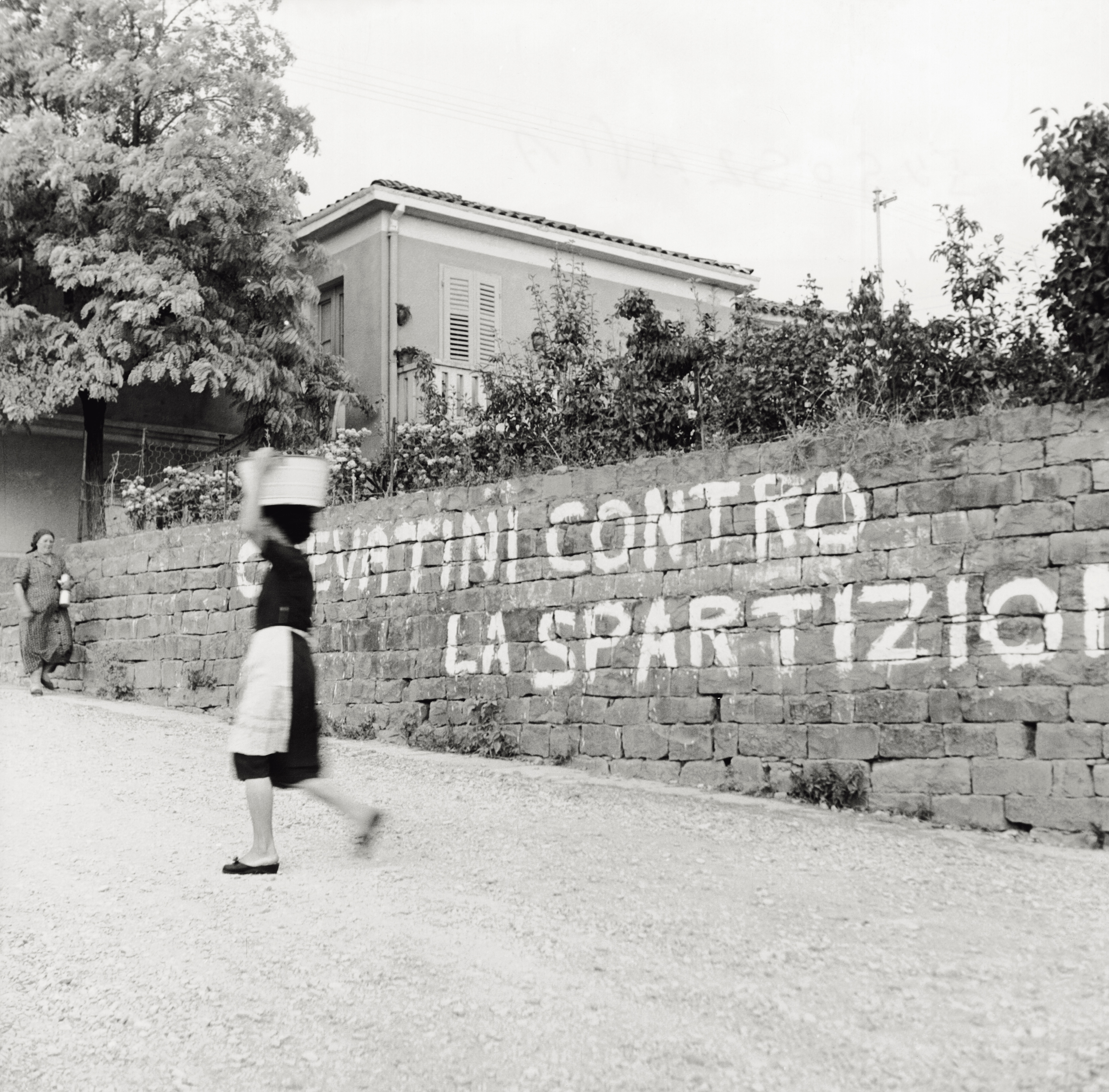 Due donne camminano su una strada di Crevattini, un paese al confine tra la zona A e la zona B di Trieste. Su un muro: 'Crevattini contro la spartizione'. La foto risale al 1954